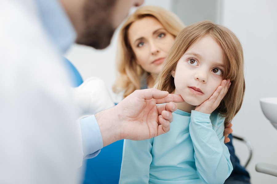 The pain is here. Worried honest cute girl showing where her tooth aching while paying a visit to the dentist with her mom