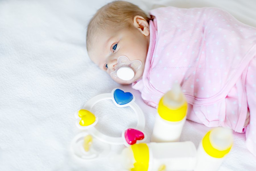 Cute newborn baby girl with nursing bottles, rattle and pacifier. Formula drink for babies. New born child, little girl laying in bed. Family, new life, childhood, beginning, bottle-feeding concept.