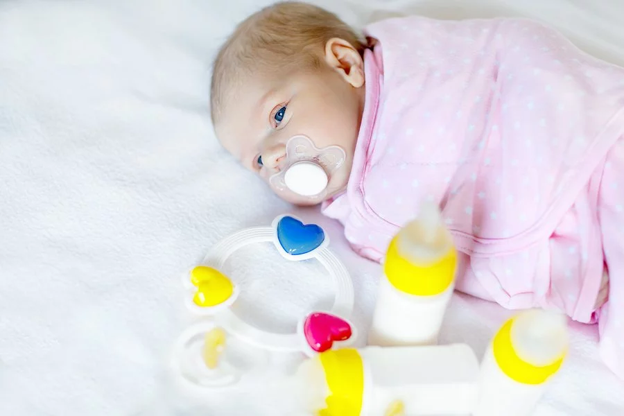 Cute newborn baby girl with nursing bottles, rattle and pacifier. Formula drink for babies. New born child, little girl laying in bed. Family, new life, childhood, beginning, bottle-feeding concept.