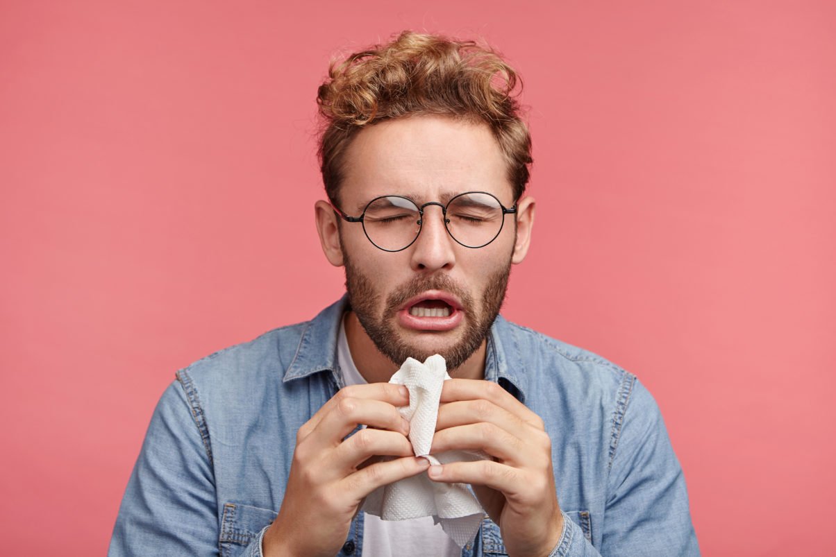 Portrait of ill or sick bearded male sneezes and coughs uses handkerchief rubs nose being allergic to animals. Man has running nose caught cold when had walk outdoors in winter or autumn