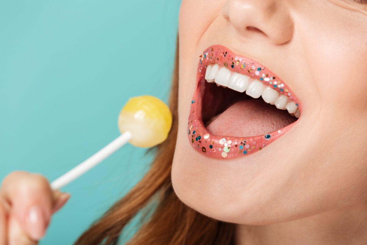 Closeup view of woman with pink lipstick with glitter on her lips and holding a yellow lollipop near her mouth.